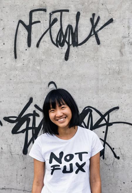 a candid amateur photograph of a young East Asian woman standing in front of a concrete wall with graffiti on it. She is wearing a white t-shirt with the text "NOT FLUX" printed on it in black letters. She has shoulder-length black hair with blonde highlights and bangs and she is smiling at the camera. The photograph appears to have been taken in an outdoor setting with trees and a building visible in the distance.
