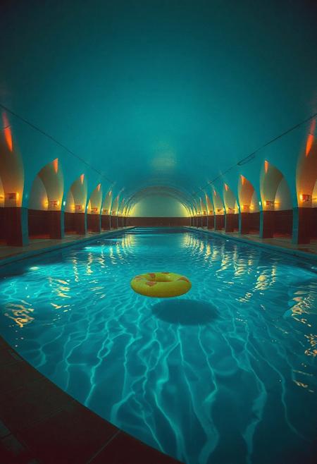 "An endless, looping pool complex with glowing teal-blue water, perfectly still yet echoing with distant, impossible splashes. The vaulted archways repeat into infinity, their neon-lit edges reflecting off the wet concrete in oily rainbow smears. The air is thick with chlorine-scented mist, distorting the flickering overhead lights. The water is crystal clear but too deep—something moves just beyond visibility, stirring the ripples unnaturally. No exits, no people—only a single abandoned pool float drifting towards the viewer. Shot on grainy 35mm film with a fisheye lens, the edges of the frame warped like a VHS tape left to decay. Vaporwave aesthetics meet psychological horror—the colors are vibrant but sickly, the geometry almost correct. Submerged shadows suggest elongated limbs just below the surface.
