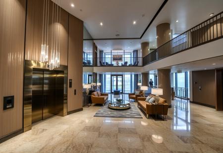 In a spacious apartment hall, there are two elevators at the front with crystal lights hanging on them. There are sofas, chairs, and tables, and the second floor corridor has metal railings visible