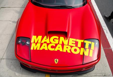 High angular shot of a 1999 ferrari 550 Maranello on a racing pit lane. It has some racing modifications. On the bonet we can see a big logo that says "MAGNETTI MACARONI" with yellow letters on two lines