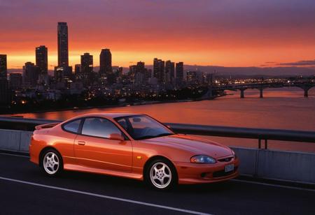 (1996 Hyundai Tiburon RD) motor vehicle, no humans, city, sky, outdoors, scenery, orange sky, road, building, cityscape, sunset, vehicle focus, skyscraper, sports car, shadow, gradient sky, red sky, evening, cloud, street, skyline, twilight, spoiler \(automobile\), orange background, dusk, red background, water, solo, bridge,