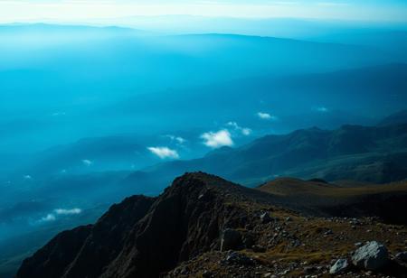 Immerse in a breathtakingly beautiful landscape: a vibrant blue-hued horizon stretches across the frame, fading into an atmospheric haze. Unusual dark colors, such as deep indigo and rich charcoal, punctuate the scene with dramatic flair. Cinematic lighting casts long shadows, emphasizing the fine details of the terrain. Film grain adds texture to the image, while superb composition guides the viewer's eye through the masterpiece. Maximum detail is achieved in every element, from the rugged terrain to the wispy clouds, creating an unparalleled level of visual mastery.