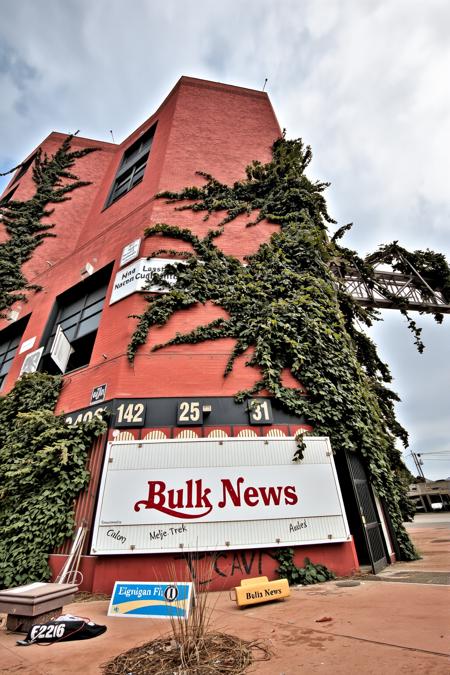 A front view of a building, Viewed from an upward looking low angle to increase its sense of height. A large abandoned brick skyscraper extends high up. The building is overgrown and infested with gigantic oversized vines with thorns. A faded and aged sign on the building reads: "Bulk News", the surrounding environment is an arid desert like environment. The scene is depicted in a photo-realistic style, There is an extreme amount of oversized vines. The window glass is broken and missing. The windows are broken and boarded over in places.<lora:Abandonedfluxv2:0.8>, abandoned