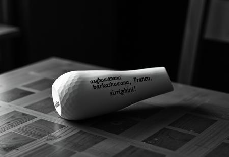 un bicchiere fatto a mano in un materiale molto lucido, che ha una resistenza enorme ma si fa modellare. ha la forma di un fungo, smussato e inclinato di 30 gradi. reca un incisione in ligua maya che suona come "asghawanana barkashawana Franco, sirrighini!" , posa su di un tavolo di legno (assi di legno spessi, vecchi e segnati dal tempo). fotografia curata da un dop professionista, noir.