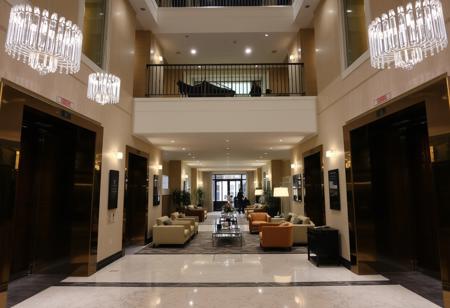 In a spacious apartment hall, there are two elevators at the front with crystal lights hanging on them. There are sofas, chairs, and tables, and the second floor corridor has metal railings visible
