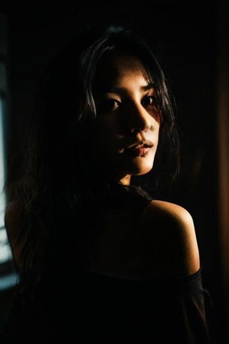 Capture the essence of a dramatic and visually striking portrait shot in a mesmerizing urban setting. Utilize a combination of lighting techniques including rim light, contour light, key light, and off-shoulder lighting to create depth and dimension. Experiment with equipment and modifiers such as beauty dish lighting, backlighting, and monochrome to enhance the mood and atmosphere. Set the scene against a dark, black background, contrasting with vibrant neon city lights visible through large windows. Embrace the surreal aesthetic of hyperkraximalism by seamlessly integrating flowing elements reminiscent of water, leaves, or clouds into the composition. Play with lighting styles like low key and high key lighting to evoke a sense of mystery and intrigue. Describe the subject with black hair and bare shoulders standing in the shadow, bathed in dim neon-colored light from blacked-out windows. The result should be a captivating blend of realism and surrealism, capturing the subject's calm expression amidst the urban energy of the cityscape.