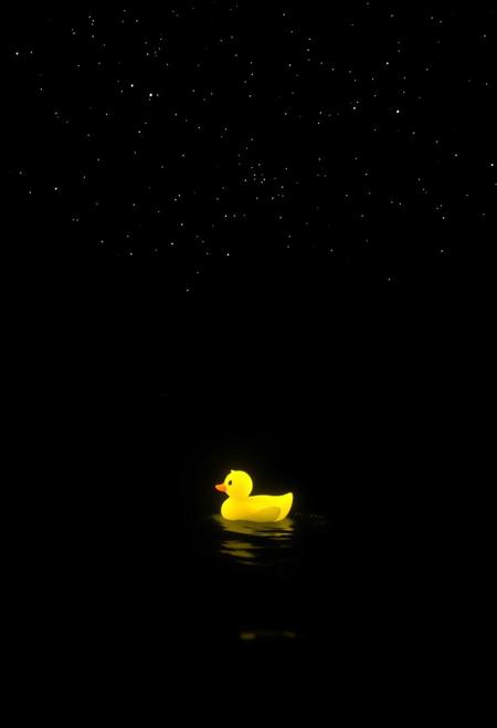 A glowing yellow duck radiates soft light as it drifts on a pitch-black sea under a starry night sky. The dark waves shimmer faintly with the duck’s luminescence, casting eerie reflections. Scattered stars twinkle above, contrasting the inky depths, evoking a mystical and tranquil atmosphere.