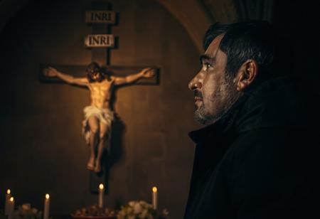 professional photo, side view photo of Caucasian man wearing a dark blue parka with dark features and a serious contemplative expression looking up at a plaster statue of Jesus on the cross mounted on the wall of a church, a sign that says "INRI" in capital letters is above Jesus on the cross, the man and Jesus are shown in profile, the scene is very dark and underexposed with only the man's and Jesus' face lit by warm, dim candle light, everything else is dark shadows