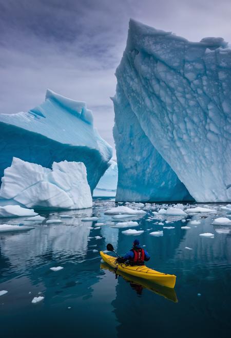 Raw photo, kayaking in Greenland: picture a lone kayaker amidst the icebergs of Greenland's icy waters. the photograph should contrast the deep blues and whites of the ice and water with the vibrant kayak, capturing the scale and isolation of the Arctic environment,