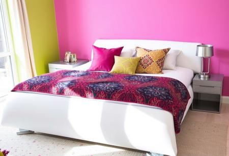 A stylish bed with sleek, contemporary design, adorned with colorful throw pillows and a vibrant patterned blanket in a modern bedroom with brightly painted walls. Next to the bed is a modern nightstand with a minimalist lamp. The space is flooded with natural light.