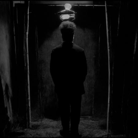 <lora:Eraserhead_Style:1> 3h3dstyl, Grayscale, black and white surreal room, inspired by Eraserhead, slightly illuminated eerie atmosphere, Henry Spencer standing with his back to the viewer, tall hair silhouette, vintage suit, dim industrial room with flickering bulb, cracked walls, warped furniture, film grain, analog horror aesthetic, subtle dust in air, high contrast shadows