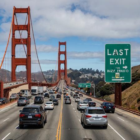 This photograph captures a prominent green highway sign with text "LAST EXIT FOR BUZZ" on a sunny day. The background shows an urban road leading to the Golden Gate Bridge, with the iconic red-orange suspension bridge towering over a busy four-lane highway. Vehicles, including a silver SUV and a blue sedan, are driving on the right lane. In the background, the cityscape of San Francisco is visible with hills and buildings under a partly cloudy sky. The image conveys a bustling urban environment, and the highway sign dominates the scene.