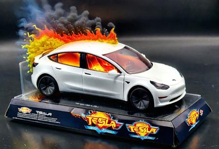Hot Wheels toy car company have made a toy car version of a Tesla Model 3, The toy telsa model 3 car comes in a mini display unit/box. The car is exploding into fire and flames, white smoke fills the top of the display box, (realistic explosion from car), (realistic Flames bellow from the car windows inside the display unit),  (standing next to the car is a little palstic toy man, he holds both arms in the air,