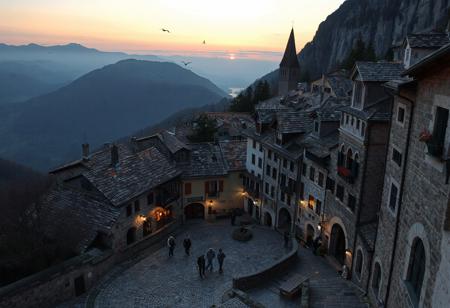 professional photo, photo of an old European mountain village with medieval cobblestone streets, a few people dot the streets but are too far to make out any details, a small flock of birds fly in the distances, no cars on the streets, at twilight, sun glows just over the horizon