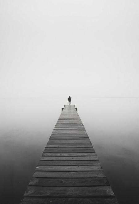 A solitary wooden pier stretches far into a motionless lake, barely visible through a veil of thick mist. The water appears like polished glass due to a long exposure effect, blending seamlessly into a soft, blurred horizon that erases the boundary between sky and water. A lone figure stands at the end of the pier, their silhouette small and dark against the expansive negative space. The entire scene is rendered in high-contrast monochrome—deep blacks and brilliant whites forming a stark visual poem. The atmosphere is cinematic and contemplative, inviting silence and introspection, like a visual haiku suspended in time. The mist blurs all detail beyond the immediate, reinforcing a dreamlike state of suspended reality.