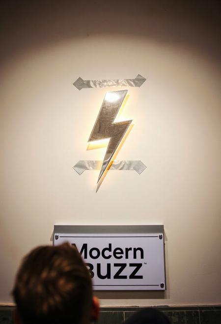 Realistic scene. Litterally a lightning bolt shaped icon that looks 3d and is glowing, duck-taped over to a blank white wall. The lightning bolt is ducked taped over only once and is about to fall off, like someone rushed to put this up. A wall sign below saying "Modern Buzz". POV is from over the shoulder of a guy, who looks confused, tilting his head and scratching his head.