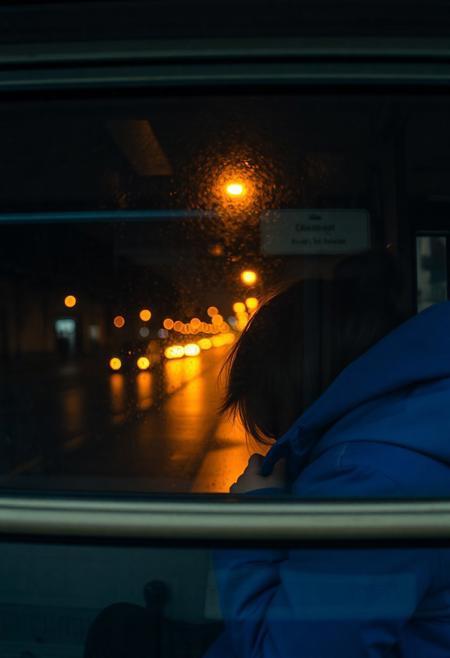 Inside an old bus, windows fogged up. Outside, dark wet streets shimmer with distorted orange streetlight reflections. A person in a royal blue coat rests their forehead against the glass. The camera zooms out through the window — the world outside feels cold, but alive.