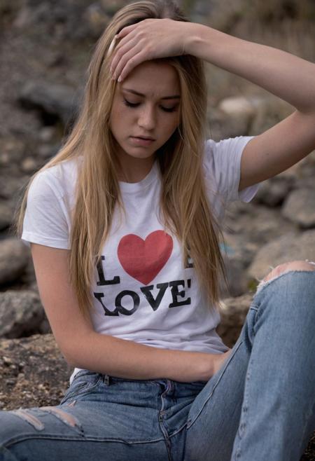 Young woman, outdoors, contemplative pose, long blonde hair, casual clothing, white t-shirt with text "LOVE", blue jeans, natural lighting, shallow depth of field, bokeh background, overcast sky, moody atmosphere, distressed jeans, fashion, stylish, hand on forehead, seated, candid expression, daytime, gray rocky ground, Fujifilm XT3, Canon R5, Fujicolor Fujichrome Velvia 100