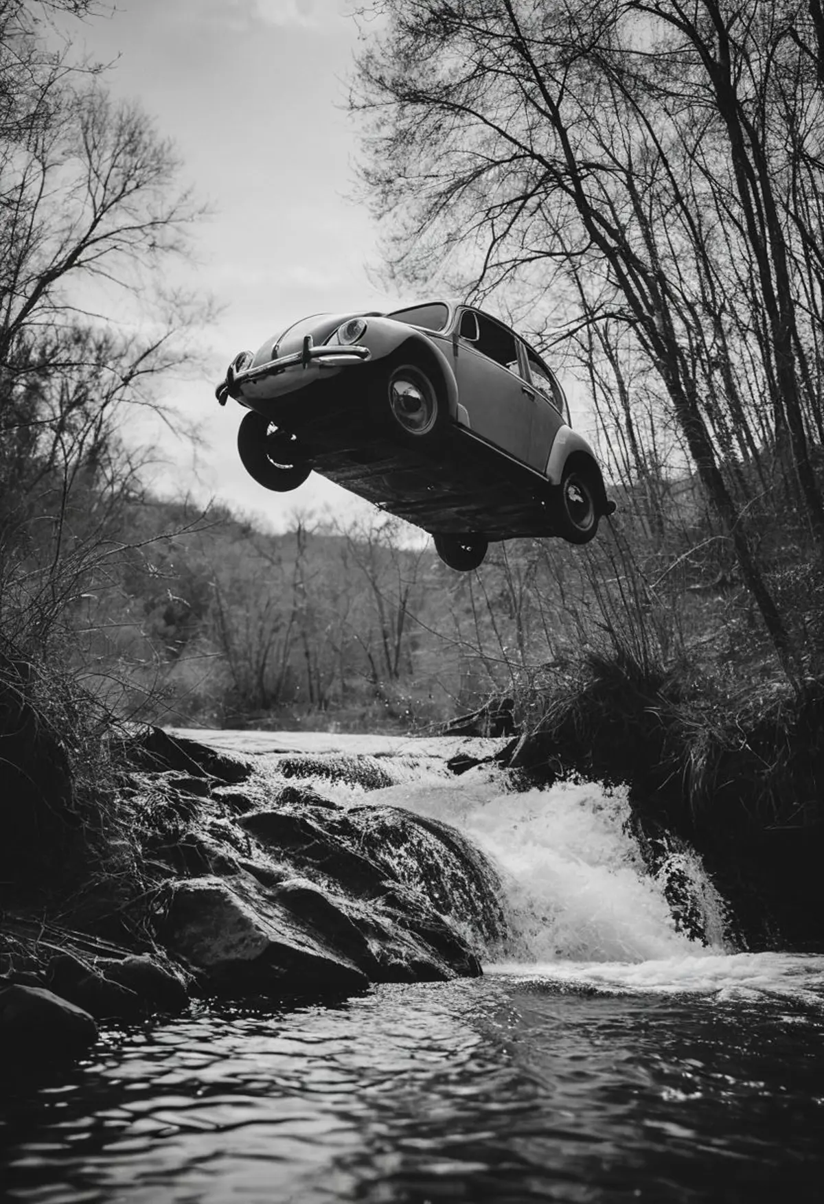A monochrome scene of a classic car seemingly suspended in midair while jumping over a rocky creek. The surrounding landscape is filled with leafless trees, suggesting a late autumn or winter season. 