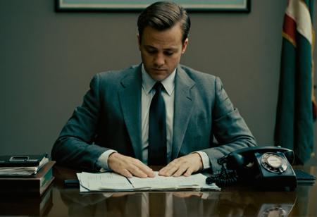 davidfincherstyle, the man is wearing a suit and tie, sitting at a desk with papers and a telephone. The man has short hair and is looking down at the papers in front of him. The room is dimly lit, creating a moody atmosphere. The image is in color, with a cool tint to it.