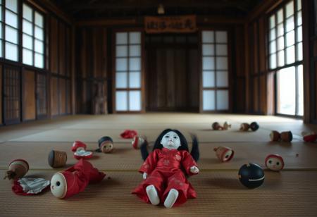 In a Japanese wooden building where a craftsman makes doll dolls, there is no one on the ground. The floor is tatami mats and the door is a horizontal sliding door. Several broken Japanese doll dolls are on the ground, including a complete doll that is 20 centimeters long. The doll is wearing a red kimono, with a pale face and long black hair in the style of a hime. Its dull expression is terrifying.