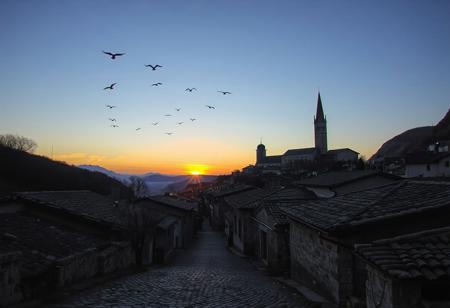 professional photo, photo of a distant old European mountain village with medieval cobblestone streets and an old cathedral, a small flock of birds fly over the town mere dots in the distance, at twilight, sun glows just over the horizon