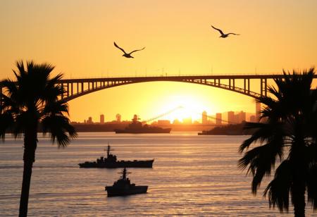 A beautiful sunrise in San Diego, California, with the warm golden light reflecting off the calm waters of San Diego Bay. The skyline is silhouetted against the vibrant hues of the morning sky, with the iconic Coronado Bridge arching gracefully across the scene. In the foreground, the historic ships of the USS Midway Museum are faintly visible, while palm trees gently sway in the soft breeze. Seagulls soar through the air, adding to the tranquil and picturesque atmosphere of the awakening city.