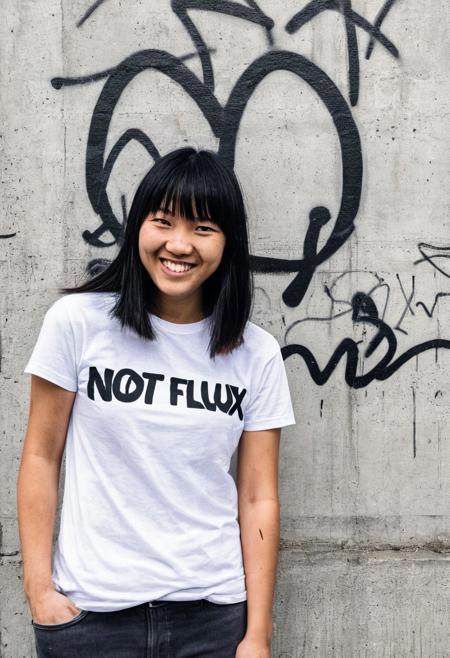 a candid amateur photograph of a young East Asian woman standing in front of a concrete wall with graffiti on it. She is wearing a white t-shirt with the text "NOT FLUX" printed on it in black letters. She has shoulder-length black hair with blonde highlights and bangs and she is smiling at the camera. The photograph appears to have been taken in an outdoor setting with trees and a building visible in the distance.
