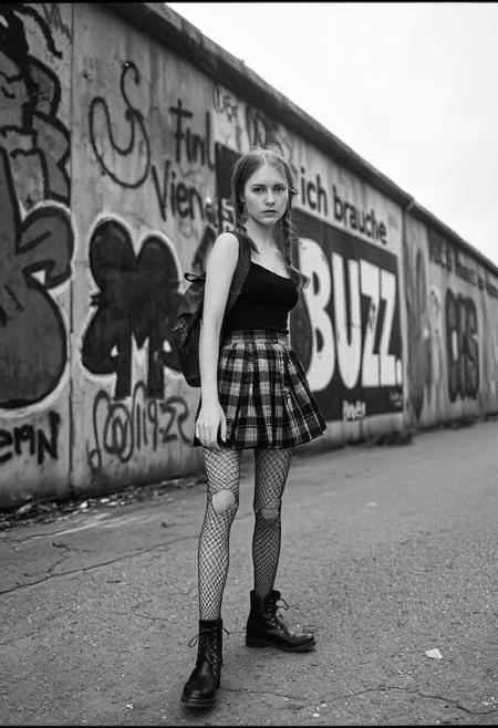 A high-contrast black and white photograph in the gritty, moody style of Anton Corbijn, shot on grainy 35mm Tri-X film. The setting is 1980s West Berlin, along the western side of the Berlin Wall. The wall is covered in chaotic, colorful German graffiti—rendered in monochrome tones—but directly behind the subject, a large, bold words “ich brauche BUZZ!!!” stands out from the rest of the markings.
In the foreground, Polly is walking along the base of the wall. She is a very slender young woman with long red hair in braids, pale freckled skin, and striking light eyes. She wears a very short black-and-grey plaid pleated skirt, a very tight black tank top, ripped fishnet stockings, and a pair of worn-in boots. A weathered leather backpack is slung casually over one shoulder.
The photo is taken from the side, capturing her stride, but her head is turned slightly toward the camera, locking eyes with the viewer. The background is stark and gritty—cracked pavement, faint fog in the air, and the towering graffiti-covered wall—but Polly is illuminated with a soft, directional light that separates her from the background. The overall mood is raw, moody, and evocative of Berlin’s underground culture in the late Cold War era. Film grain, deep shadows, and high contrast emphasize the documentary-style authenticity.