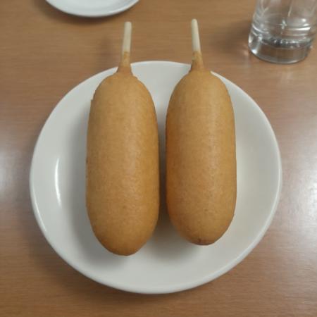 A photorealistic image of a delicious 9hotdog placed on a plate in a restaurant setting. The 9hotdog should be golden brown, with a crispy exterior and a juicy interior visible at one end where it's bitten off. The plate can be white or a light color to contrast with the golden hue of the 9hotdog. There might be a small dish of mustard or ketchup on the side for dipping. The table should have a clean tablecloth, and the lighting should be warm, highlighting the appetizing look of the 9hotdog. The background can have subtle hints of a restaurant ambiance, like a glass of water or some cutlery, <lora:cornhotdog:0.65>, (bottle of soda),<lora:applesoda:0.75>