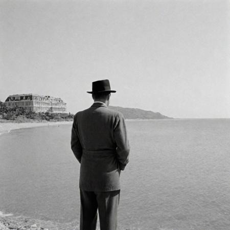 1950s detective film, lordly grandhotel standing directly at the water of the sea, surrounded by beaches and lush plantlife. Captured on a 35mm camera, black and white, classic film noir film.