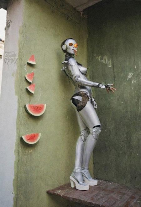 Editorial photograph, three gorgeous female brunette cyborg artists, dancing on the balcony of a typical messy, twenty-something's, So Paulo, Brazil apartment in the Liberdade barrio, a sassy but annoyed and slightly damaged gay robot stands in the corner smoking a cigarette, bad paintings of watermelon hang on wall, 1970s retrofuturism, dramatic moody cinematic lighting, Leica M3, film grain,