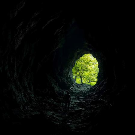 Dark, jagged cave interior with protagonist slowly advancing toward a small opening revealing lush green foliage outside, harsh contrast between shadowy confinement and bright natural world representing hope and escape.