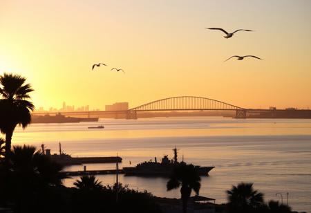A beautiful sunrise in San Diego, California, with the warm golden light reflecting off the calm waters of San Diego Bay. The skyline is silhouetted against the vibrant hues of the morning sky, with the iconic Coronado Bridge arching gracefully across the scene. In the foreground, the historic ships of the USS Midway Museum are faintly visible, while palm trees gently sway in the soft breeze. Seagulls soar through the air, adding to the tranquil and picturesque atmosphere of the awakening city.