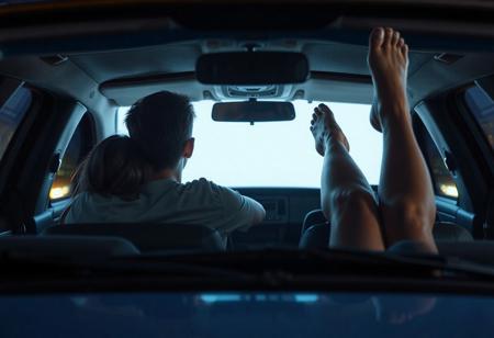 view of front of car at drive-in movie, screen lighting through the windshield dimly the front seat, where a 18yo couple have their heads leaning together, his arm around her shoulders, and  lighting the back seat where a woman's widespread bare legs are sticking up in the air, shoe hanging from one foot. pov: movie screen facing car. 
, very detailed, detailed eyes,  high detailed texture, realistic, photorealistic, perfect eyes, real life realistic,  detailed background,