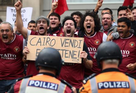 A photograph of a screaming, ragged, angry group of Torino FC fans, wearing a burgundy soccer vest, protesting with a cardboard in hands that says "CAIRO VENDI E VATTENE!", police officers stand in the foreground with their backs to the viewer,