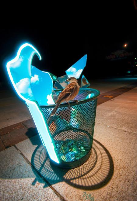 JNANX,luminous white silhouette,cyan glow,emerald highlights,flickering outline,dreamlike movement,rippling colors,drifting embers,infinite black background,psychedelic colors,ethereal,futuristic surrealism,gw,glowing,night,night sky,trippy,
Prompt: A street-level, eye-level shot of a dark green, cylindrical metal mesh trash can sitting on a concrete sidewalk. Inside the trash can, several pieces of a broken mirror reflect a bright blue sky with fluffy white clouds. A small brown sparrow with darker streaks sits perched on the rim of the trash can, looking towards the reflected sky within the mirror shards. The sunlight casts a shadow of the trash can and the bird onto the sidewalk.