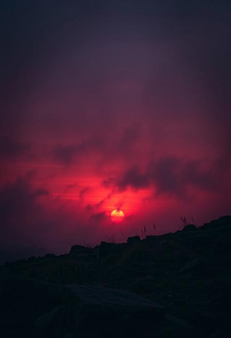 A crimson-hued sunset descends upon a rugged terrain, where wispy clouds and misty fog envelop the landscape in an eerie atmosphere. A dark, muted palette dominates, with deep oranges and purples bleeding into the distant haze. Cinematic lighting casts long shadows, imbuing the scene with dramatic tension. The camera pans across the terrain, capturing every nuance of fine detail, from the wispy grasses to the weathered rocks. A masterpiece of composition and atmospheric mastery, this breathtaking image demands attention.