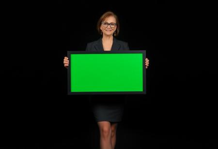 A nerdy 56yo Milf wearing a sexy business suit, short hemline, sheer pantyhose,  standing in front of a pure black background, holding up a blank (chroma greene sign) with a black border, full body view, ultra realistic, high def, HD, 8k