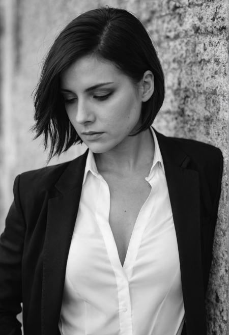 (black and white), (portrait), (closeup), (stern look), maskuline 25 years old  woman, flat breasts, black jacket, open white shirt, black tie,  (looking down at her chest), leaning at wall, short raven bob, (cinematic), bokeh,  (film grain)