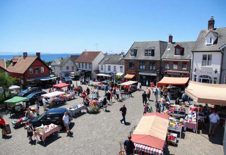 small town Saturday market day, sunny, northerly, July, crisp, near the sea,, village square filled with 40 tables, 20 booths, crafts, homemade food, carvings, buyers, sellers, colorful, chocolate sop, bakery, parked cars, produce, berries, pottery, wool weaving, hot dog grilling, bookstore, bank building, tea room, view from above