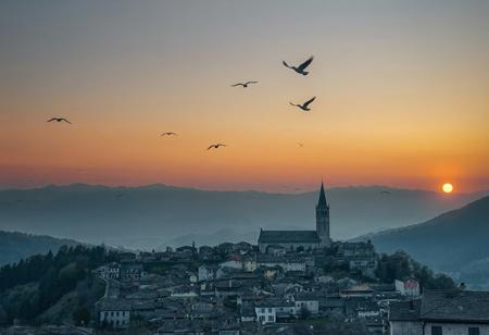 professional photo, photo of a distant old European mountain village with medieval cobblestone streets and an old cathedral, a small flock of birds fly over the town mere dots in the distance, at twilight, sun glows just over the horizon