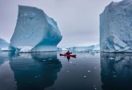Raw photo, kayaking in Greenland: picture a lone kayaker amidst the icebergs of Greenland's icy waters. the photograph should contrast the deep blues and whites of the ice and water with the vibrant kayak, capturing the scale and isolation of the Arctic environment,