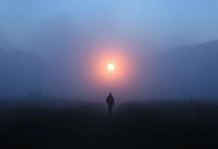Orb as a Guiding Light in a Foggy Moor: A lone traveler walks through a dense, fog-covered moor at twilight. Ahead of them, a soft, glowing orb drifts just above the ground, guiding their way through the mist. The surrounding landscape is barely visible, with only the faint outlines of distant trees and hills in the thick fog.