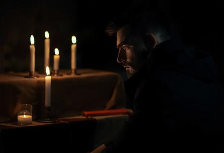 professional photo, side view photo of Caucasian man in kneeling in front of an altar with 3 lit candles on it, wearing a dark blue parka, serious face, contemplative expression, dark features, dramatic shadows, very dark and underexposed, face lit only by candles, all else dark