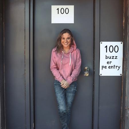 a locked door with a white sign that reads "100 buzz per entry". Outside the door there is a woman 26 years old model. Very clearly desperate to pee. Wearing jeans. Wearing pink hoodie. Squeezing legs together tightly. Crossed legs. Very desperate to pee. Door is closed. She is looking at viewer. She is smiling. She really need to pee.