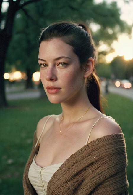 Grainy lowfi desaturated film photograph taken in a city park at dusk. Heavy Bokeh effect with focus on her green eyes, blurry background. A pretty young woman of northern Italian heritage wraps her brown cardigan around her bare shoulders. Goosebumps on a bare exposed shoulder. She wears the cardigan over a little summer sun dress and is chilly as the cool wind blows loose stands off her long black hair in her eyes. Tight ponytail. Fine Delicate thin gold chain necklace. She resembles Liv Tyler. The setting sun casts soft light into her eyes. Full soft cheeks and softer jawline, soft rounder face with the lightest freckles.