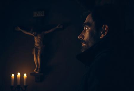professional photo, side view photo of Caucasian man wearing a dark blue parka with dark features and a serious contemplative expression looking up at a plaster statue of Jesus on the cross mounted on the wall of a church, a slightly out of focus single sign that says "INRI" in vague capital letters is above Jesus on the cross, the man and Jesus are shown in profile, the scene is very dark and underexposed with only the man's and Jesus' face lit by warm, dim candle light, everything else is dark shadows