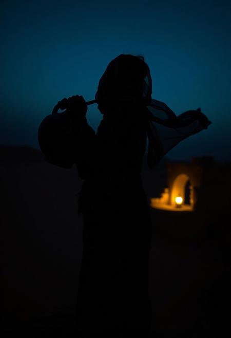 silhouette shot, a North African woman balancing a clay water jug on her shoulder, standing hip-cocked at the edge of a desert oasis, sheer scarf fluttering in the wind, looking back at the viewer with half-lidded eyes, setting: dunes and crumbling stone arches, mood: sultry survival, lighting: deep blue hour with a single lantern glow, shot on Leica M10-P, styled like a desert noir film