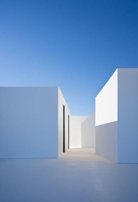 Capture the essence of minimalism through a conceptual design of a building with perfect geometry, using a white and blue color palette. Emphasize the interplay of light and shadows to highlight the clean lines and precise angles, creating a composition that exudes simplicity and elegance. Include a silhouette figure in the scene to add a sense of scale and human connection, standing between the architectural elements. Utilize soft, diffused blue lighting to enhance the crisp white surfaces and deep shadows, emphasizing the structure's architectural purity. This minimalistic approach will bring out the building's inherent beauty, showcasing the harmony between light, shadow, and form against a serene blue backdrop.
